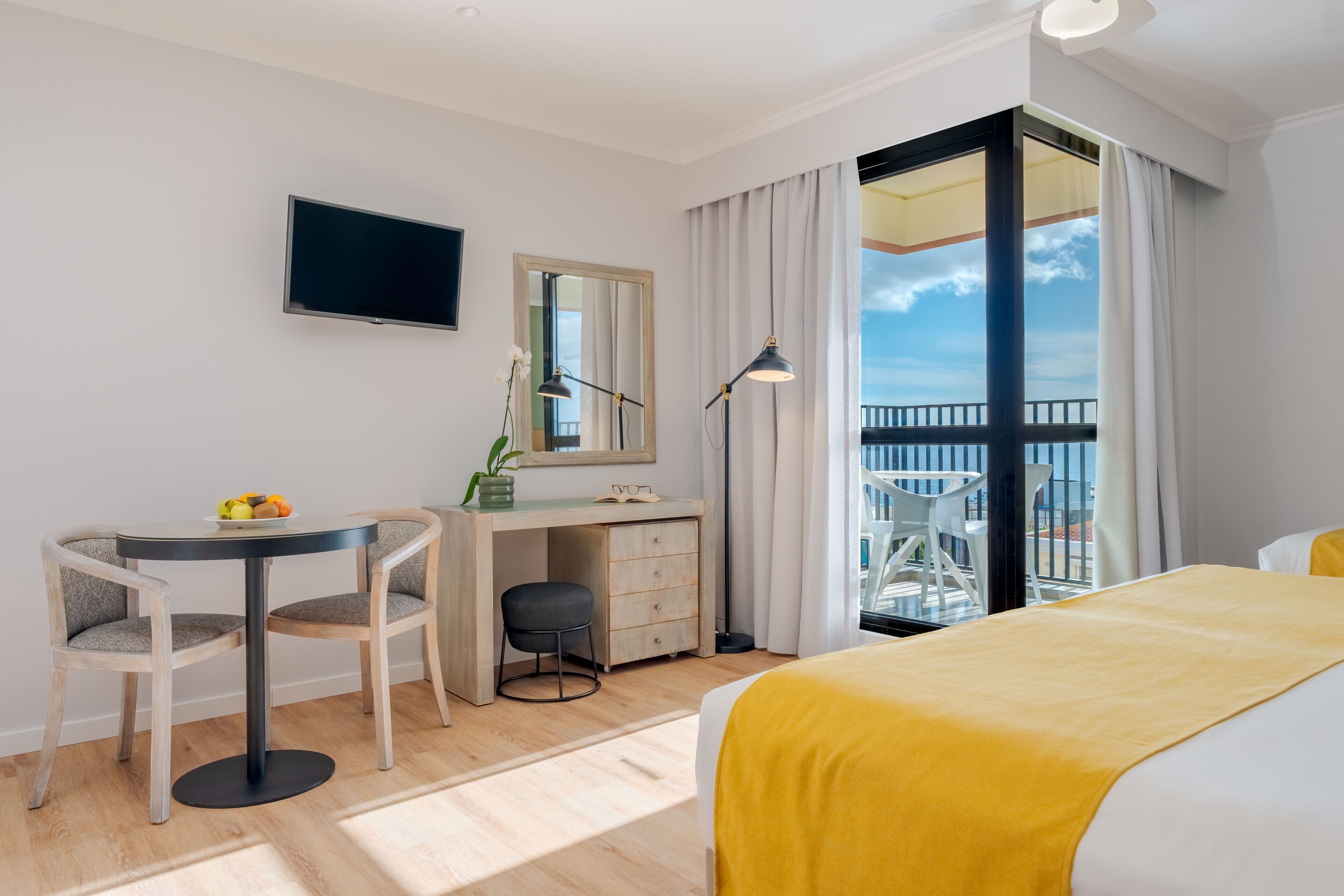 A hotel room with a bed, a desk, a chair, a TV, a balcony with a view, and a window with curtains.