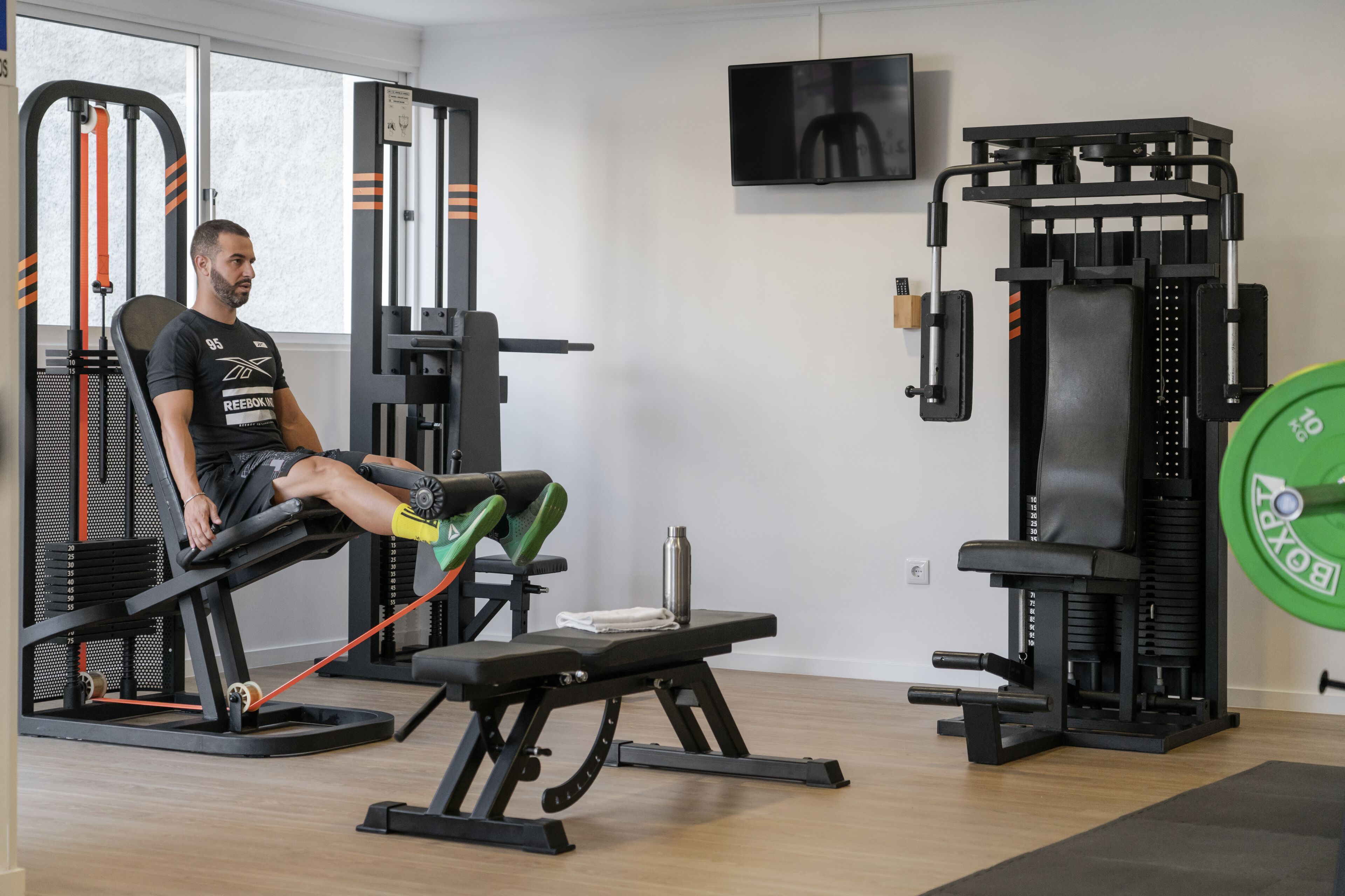 A gym with exercise equipment, a bench, and a television on the wall.