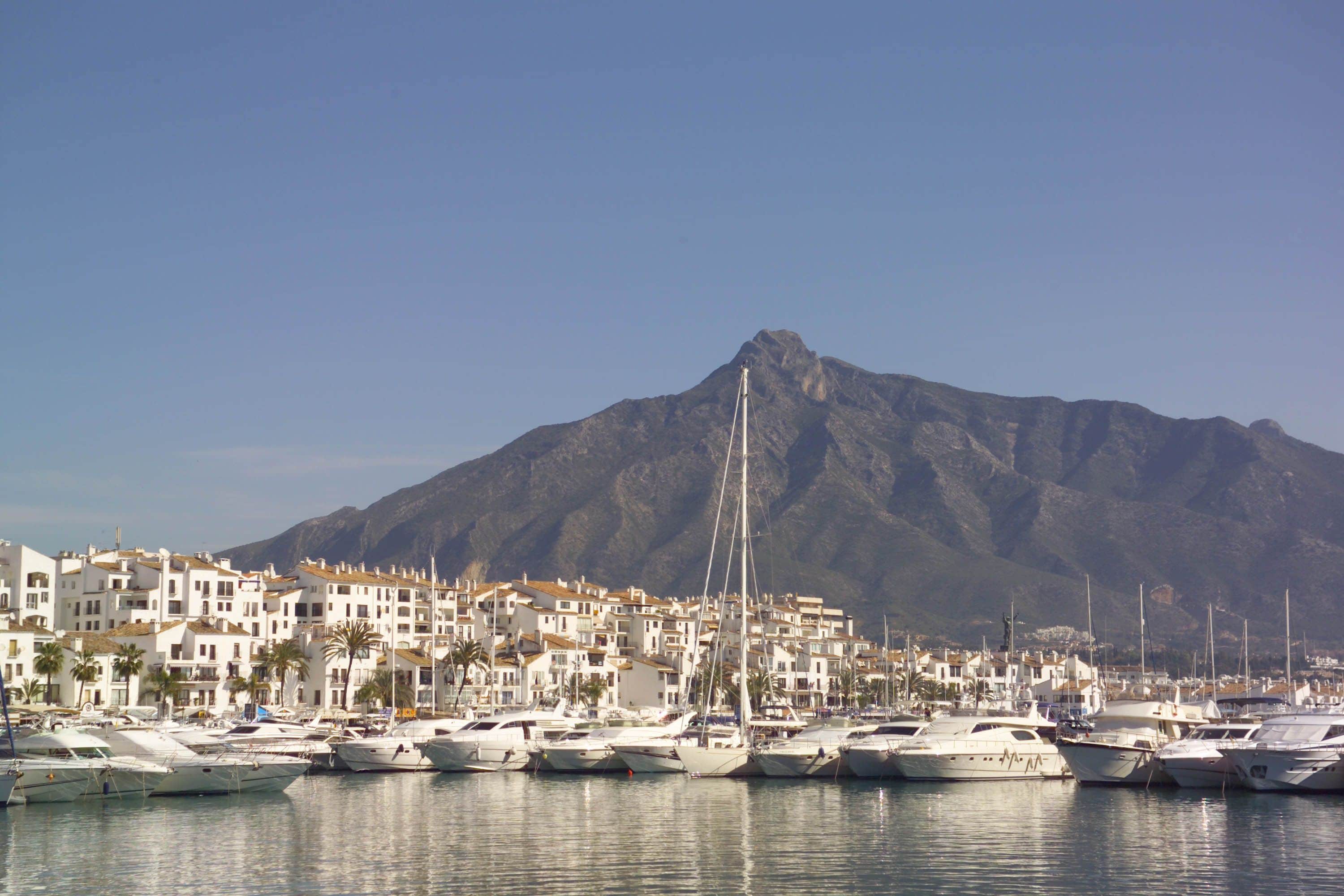 Un port de plaisance où sont amarrés des bateaux, avec une ville côtière aux bâtiments blancs en arrière-plan et une montagne qui se profile à l’horizon.