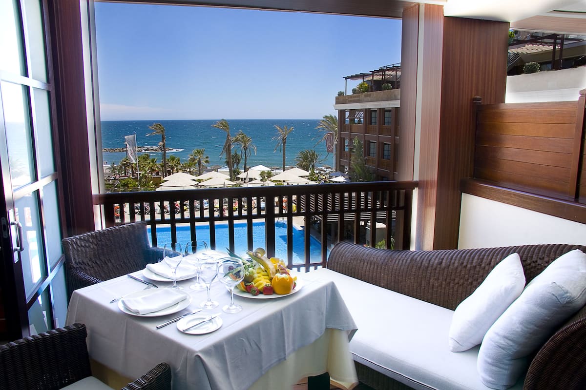 A balcony with a view of the ocean, a dining table set with plates, glasses, and a fruit bowl.