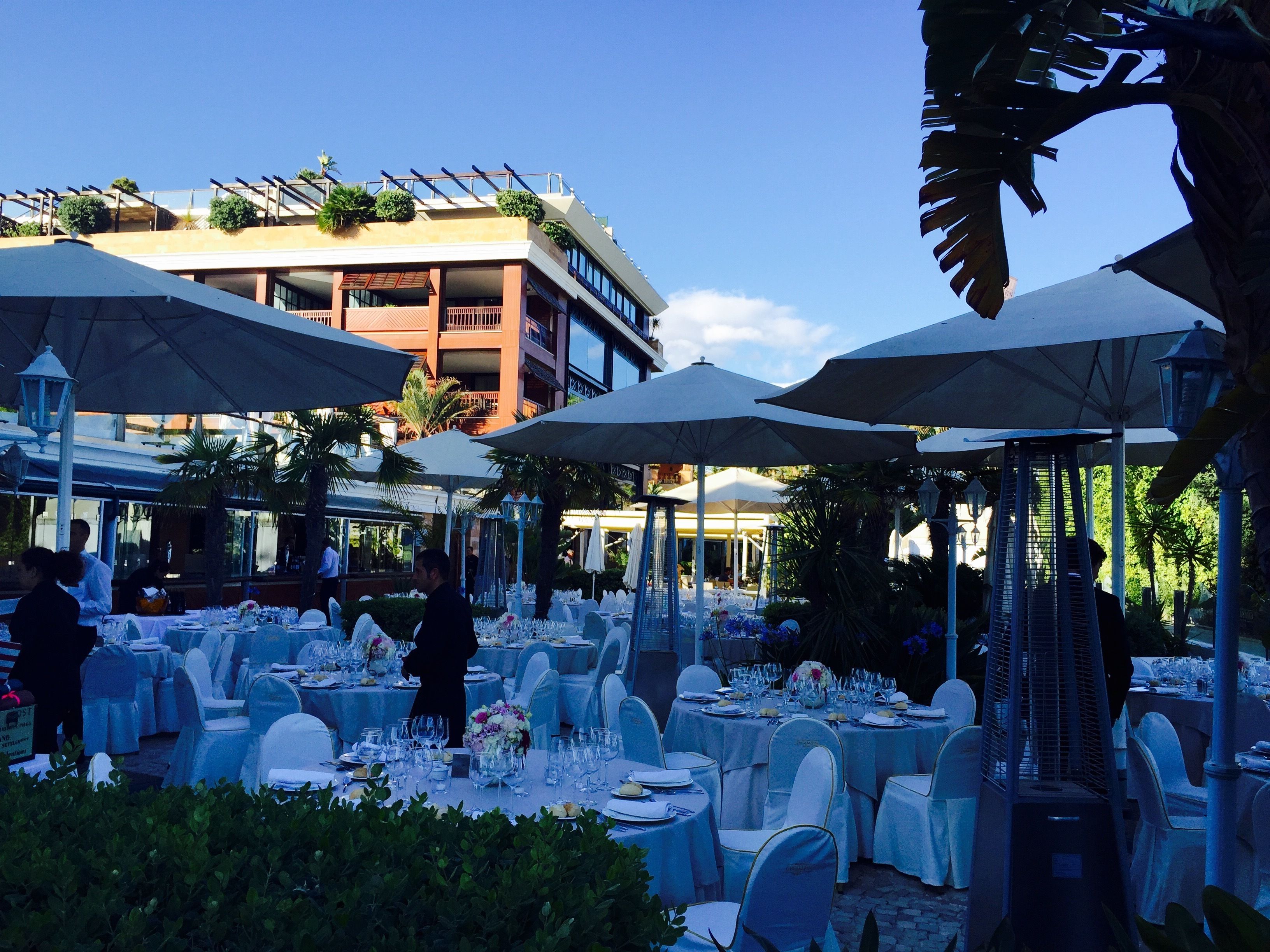 Un área de comedor al aire libre con mesas redondas dispuestas para un evento formal, rodeada de sombrillas y sillas.