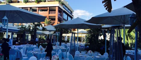 An outdoor dining area with round tables set for a formal event, surrounded by umbrellas and chairs.