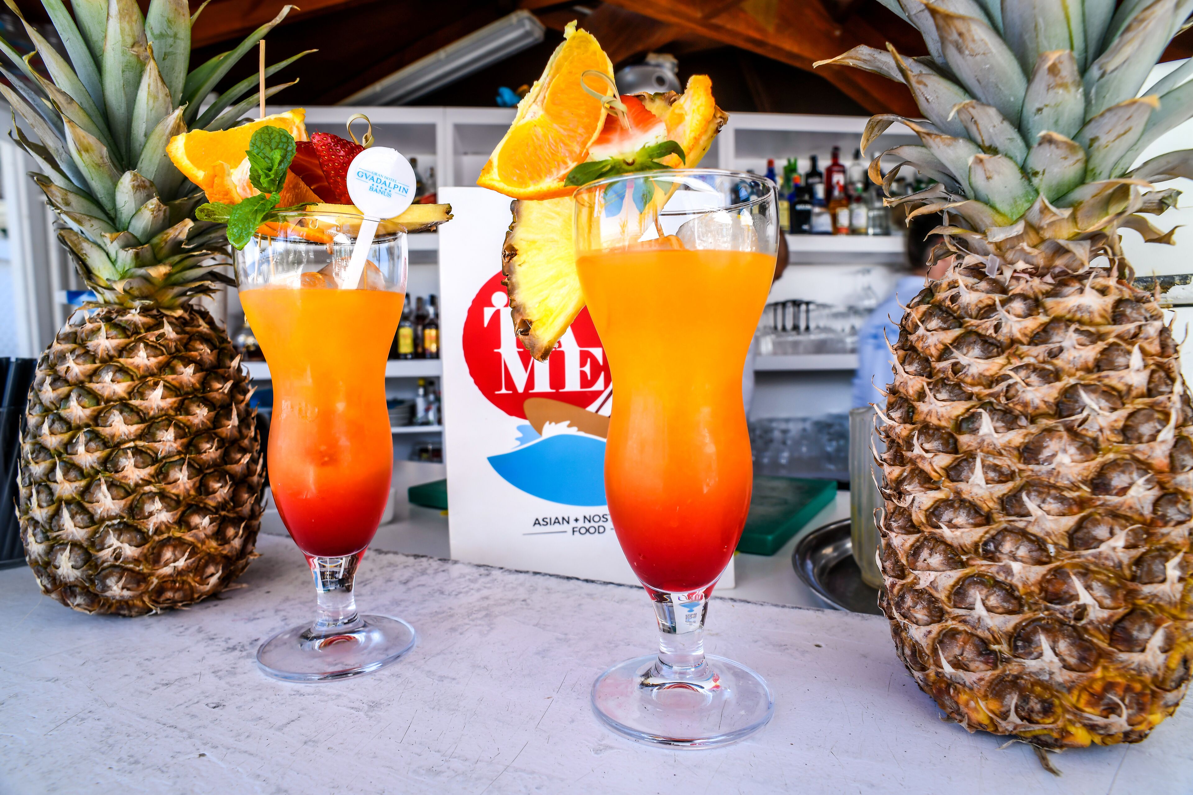 Two glasses of a red-orange beverage with pineapple garnishes and a sign in the background.