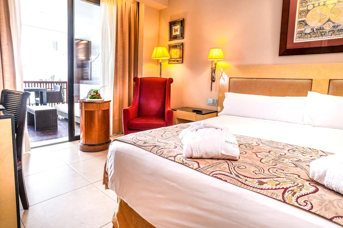 A hotel room with a large bed, a red armchair, two bedside lamps, a small table, and a view of the outside balcony.