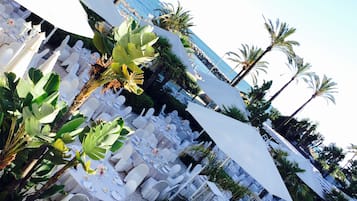 Un área de comedor al aire libre con sillas y mesas blancas, rodeada de plantas tropicales y palmeras, con vista a un cuerpo de agua.