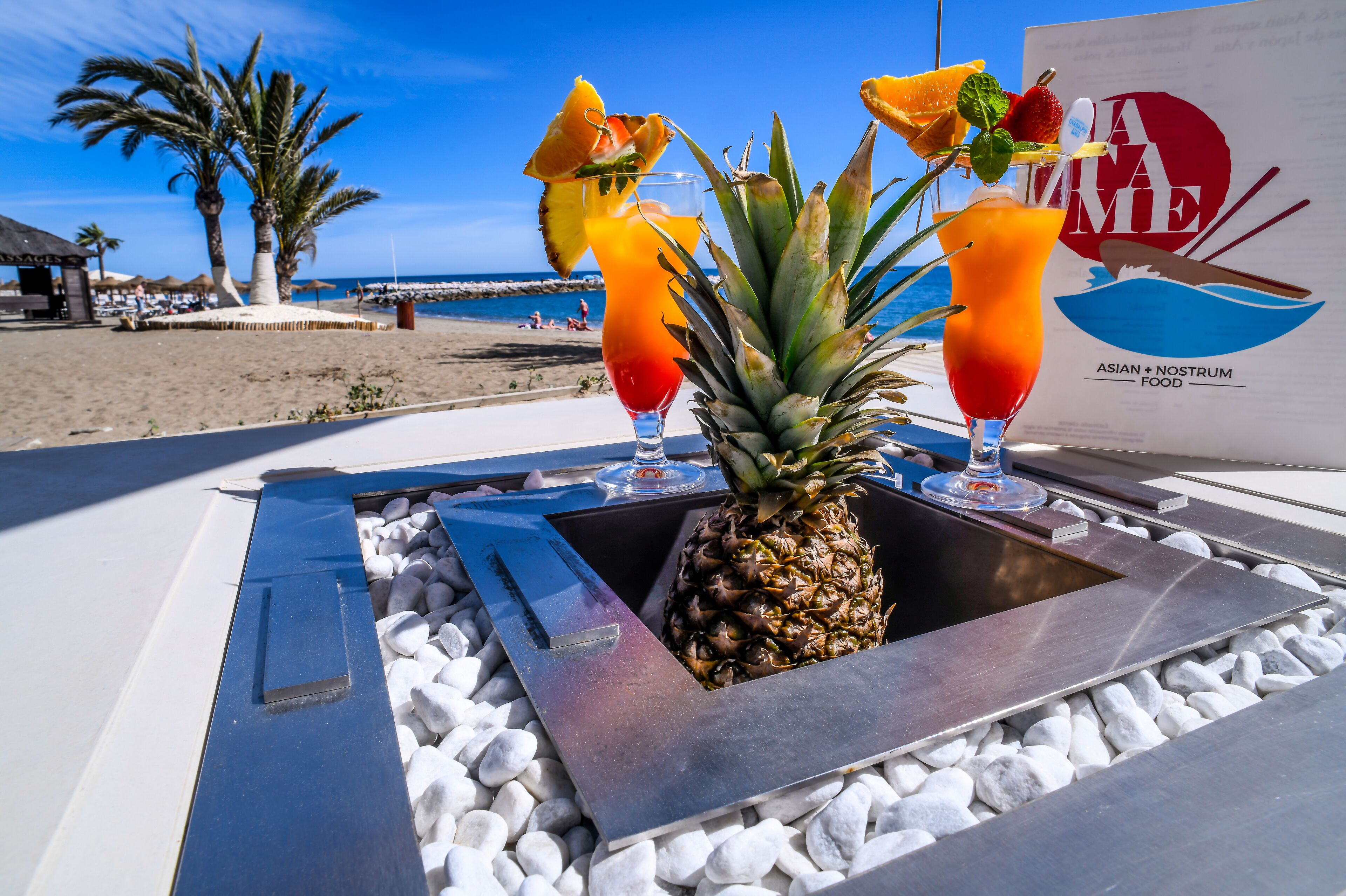 Disposición de bebidas tropicales con una piña como pieza central, vista a la playa y un letrero que anuncia comida asiática + Nostrum Food.