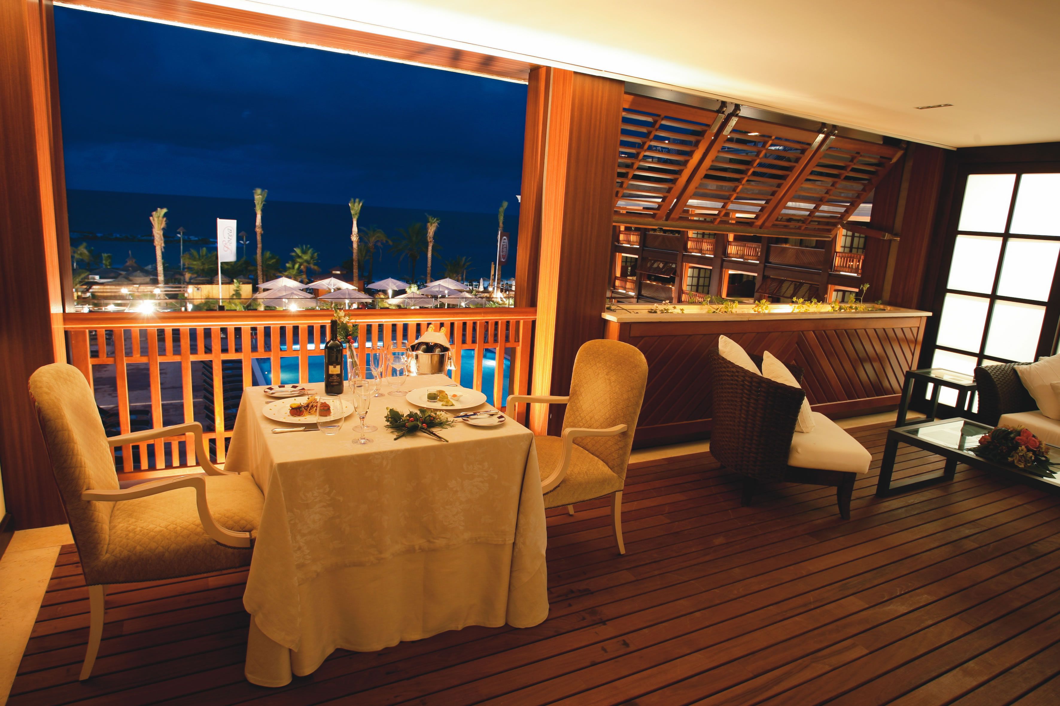 A dining area with a table set for two, overlooking a pool and palm trees.