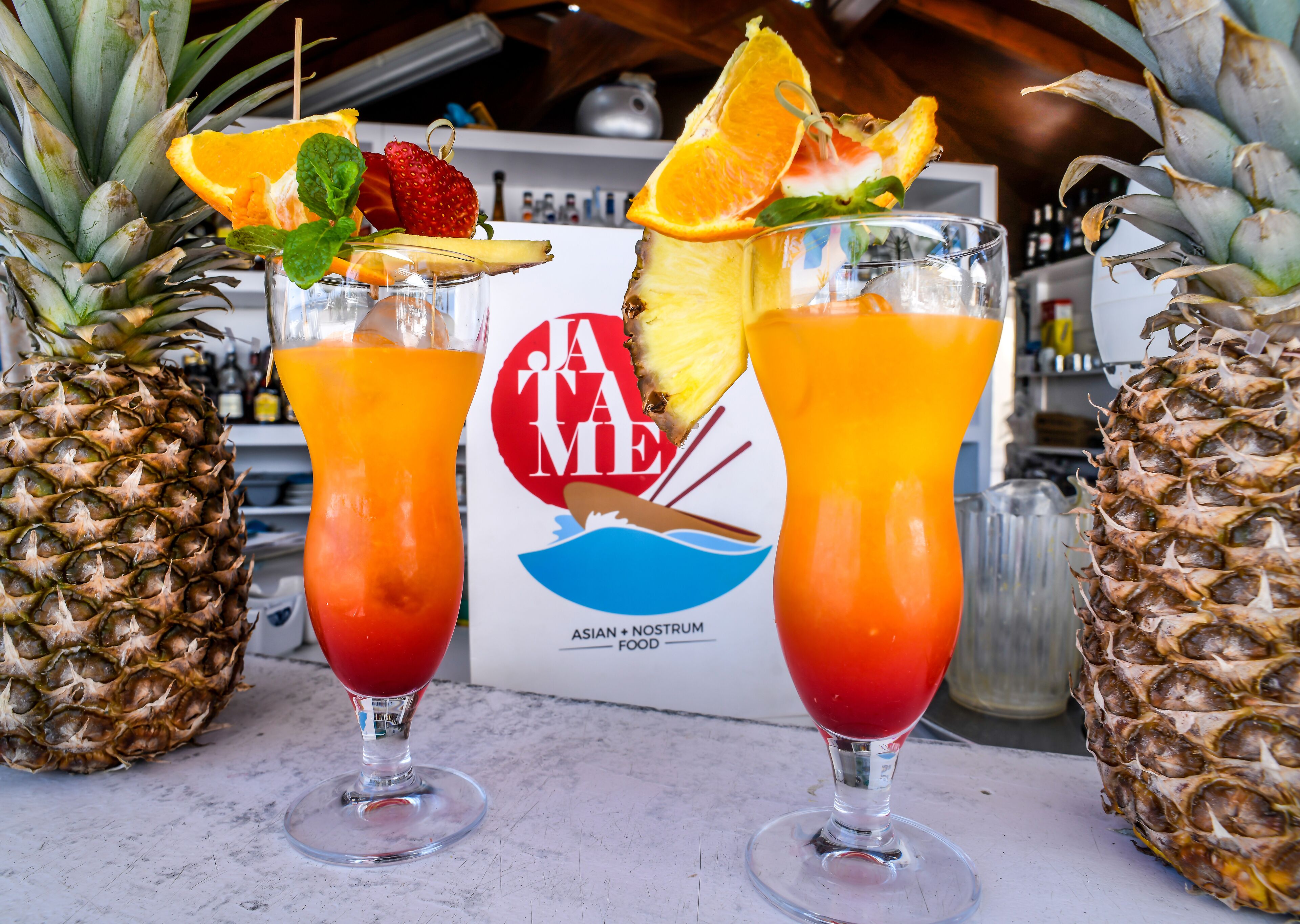 Twee cocktails, versierd met fruit en muntblaadjes, met een bordje op de achtergrond waarop staat: JAMAICA MEAT ASIAN + NOSTRUM FOOD.