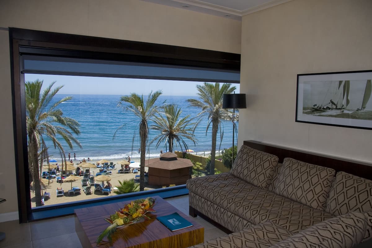 A room with a large window overlooking a beach, a patterned sofa, and a small table with a vase.