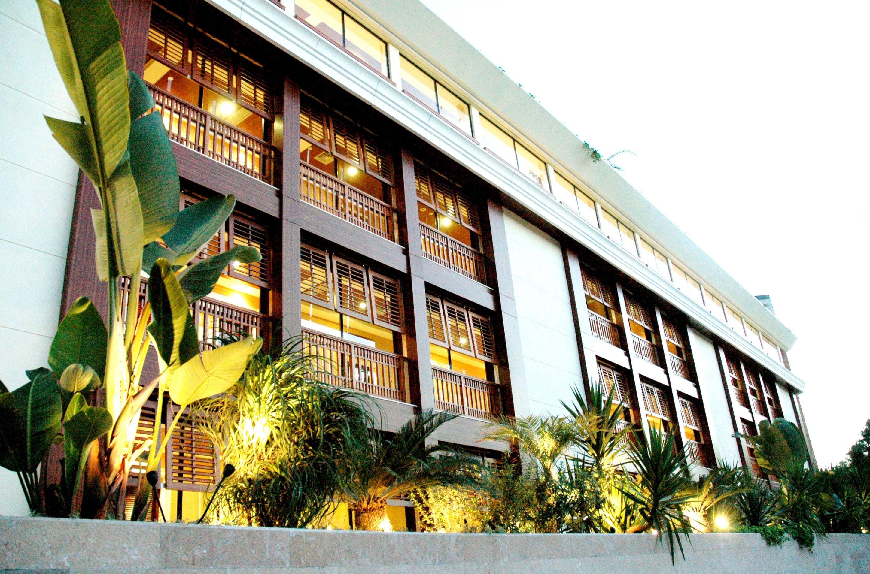 A multi-story hotel with balconies, large windows, and lush tropical plants in the foreground.
