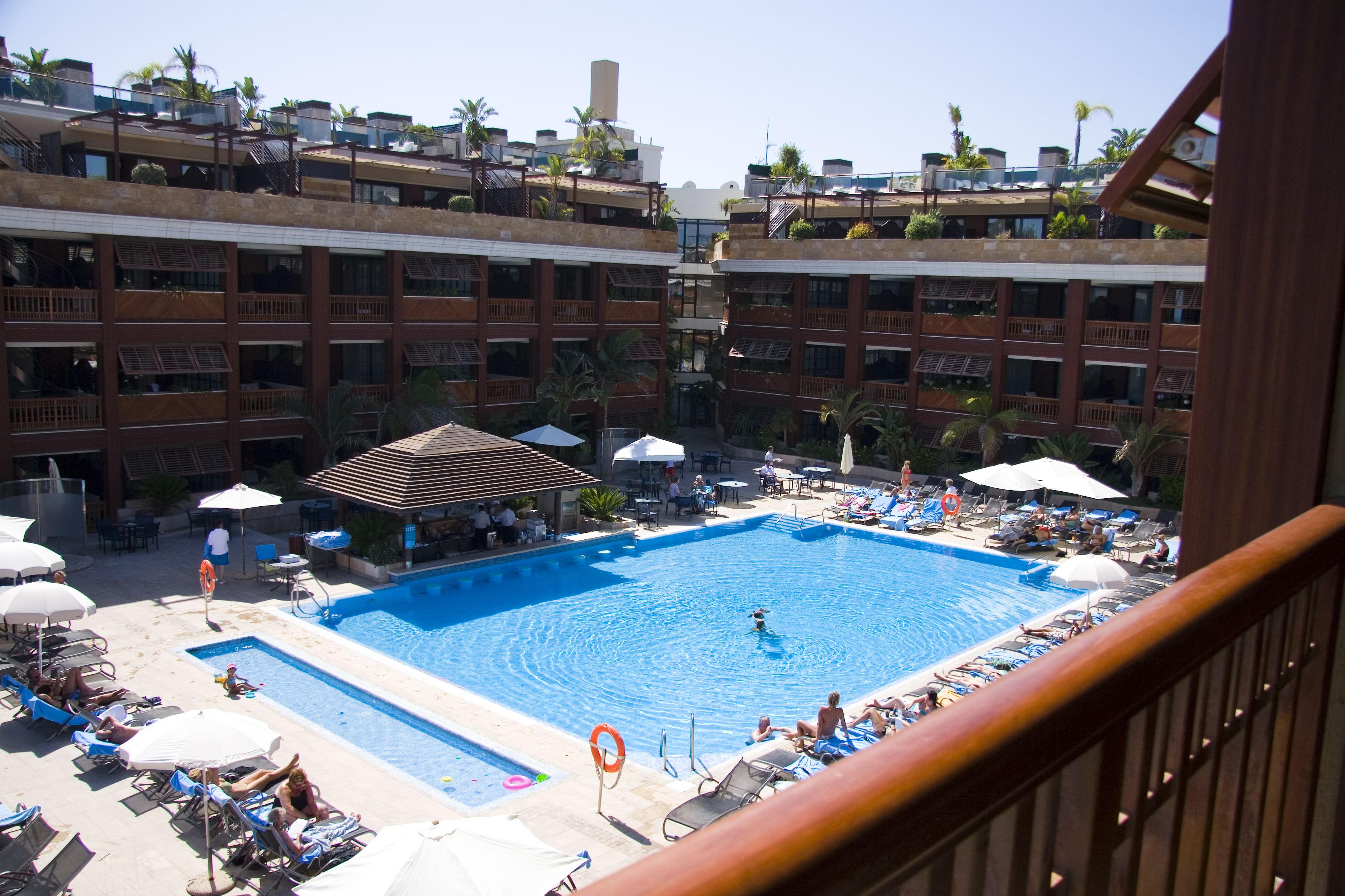 A hotel pool area with multiple swimming pools, sun loungers, umbrellas, and a covered seating area.