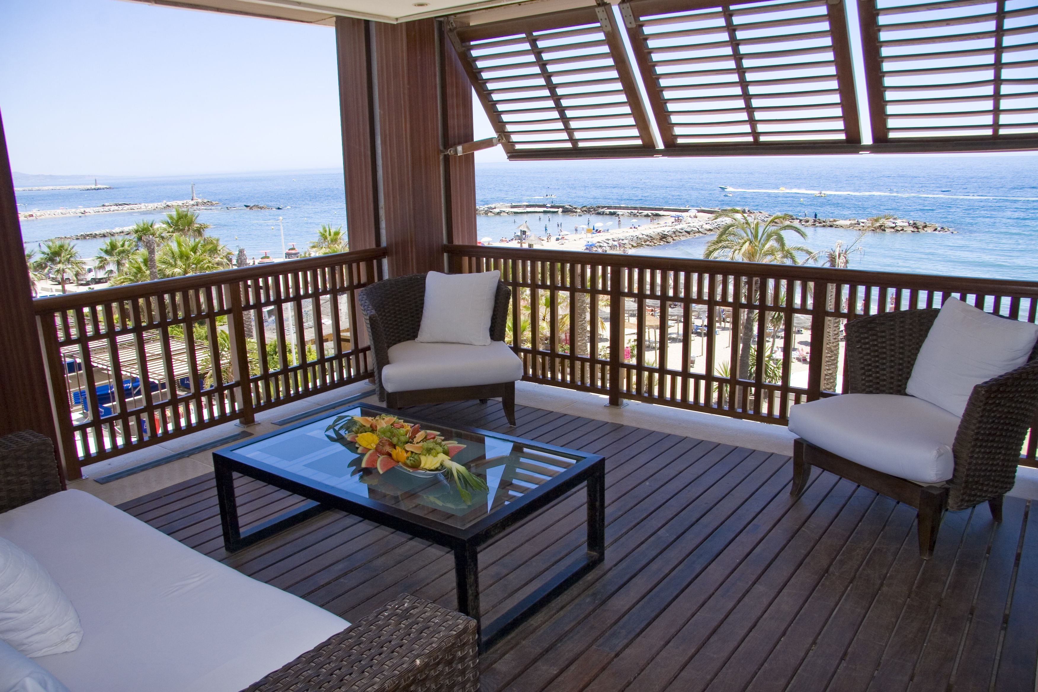 A balcony with wicker furniture, a glass table with fruit, and a view of the sea.