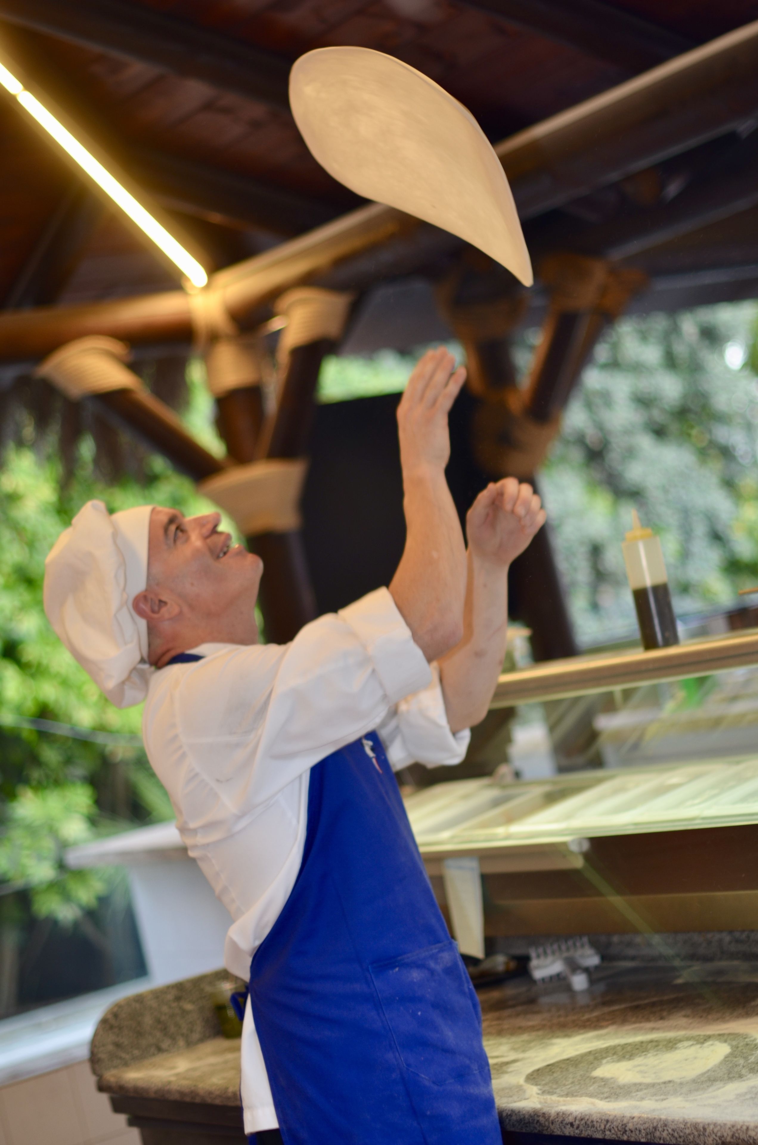 A chef in a white chef's hat and blue apron, reaching up to toss a large, round, white object.