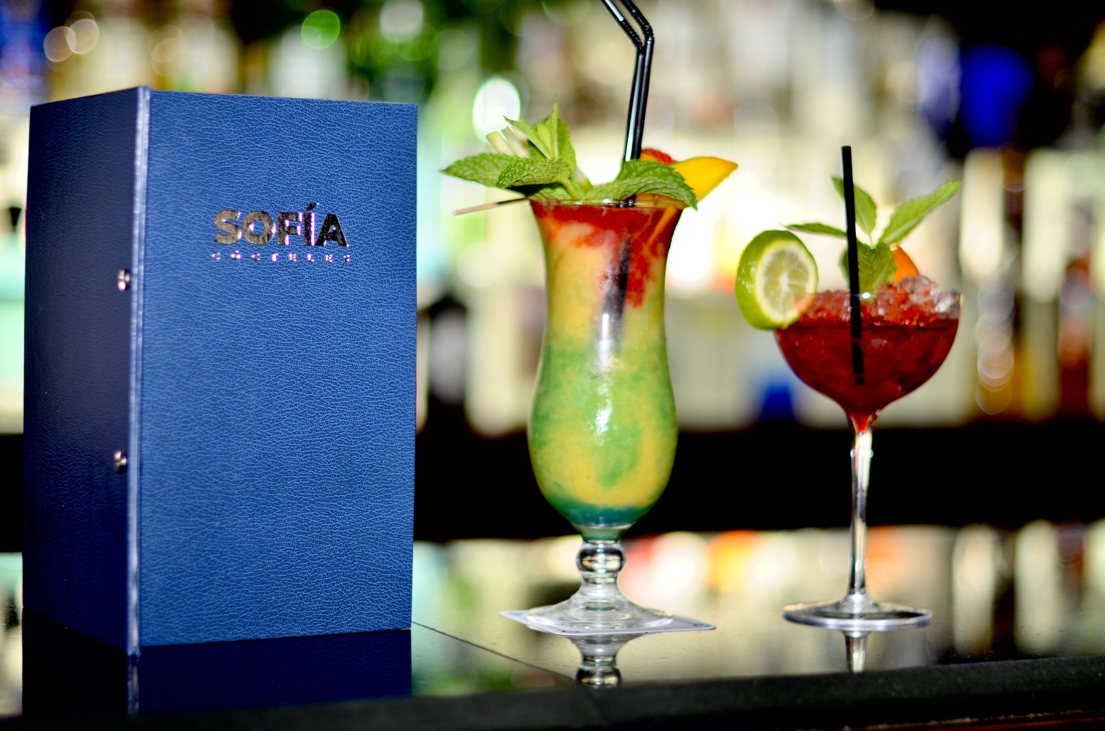 Dos cócteles en vasos con pajitas, una caja azul con el rótulo "Sofia" y un fondo desenfocado de botellas.