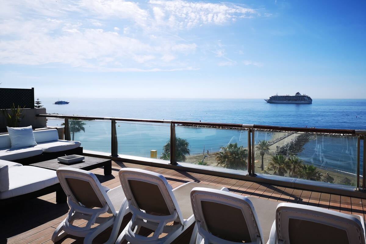 A balcony with white chairs and a sofa, overlooking a calm sea with a cruise ship in the distance.