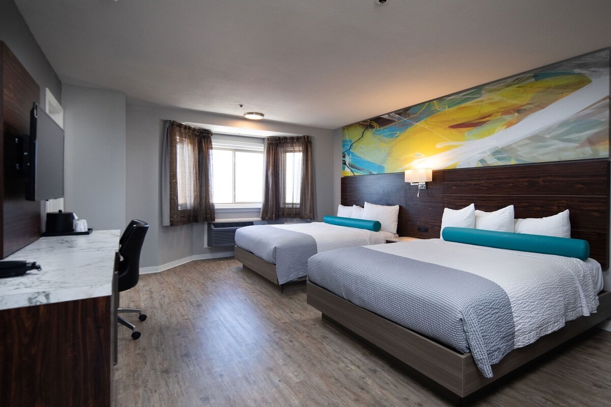 A hotel room with two beds, a desk, a chair, a window with curtains, a wall-mounted lamp, and a large abstract painting.
