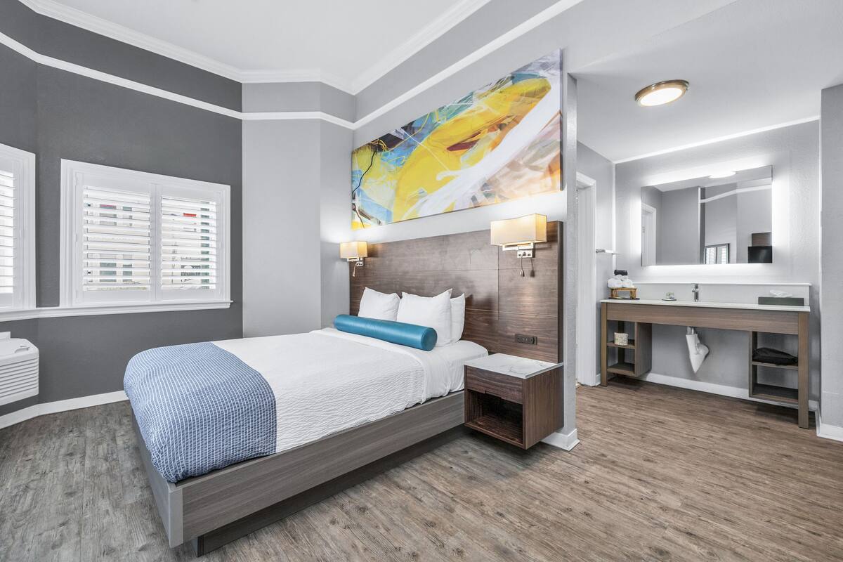 A modern hotel room with a large bed, a bedside table, a vanity with a mirror, and a large abstract painting on the wall.