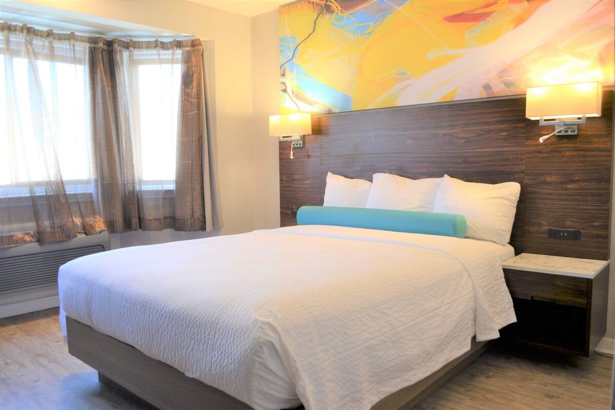A hotel room with a large bed, a nightstand, a wall-mounted lamp, and a window with curtains.