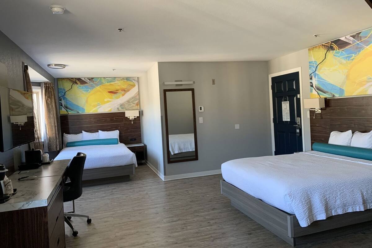A hotel room with two beds, a desk, a chair, a large mirror, and a door.