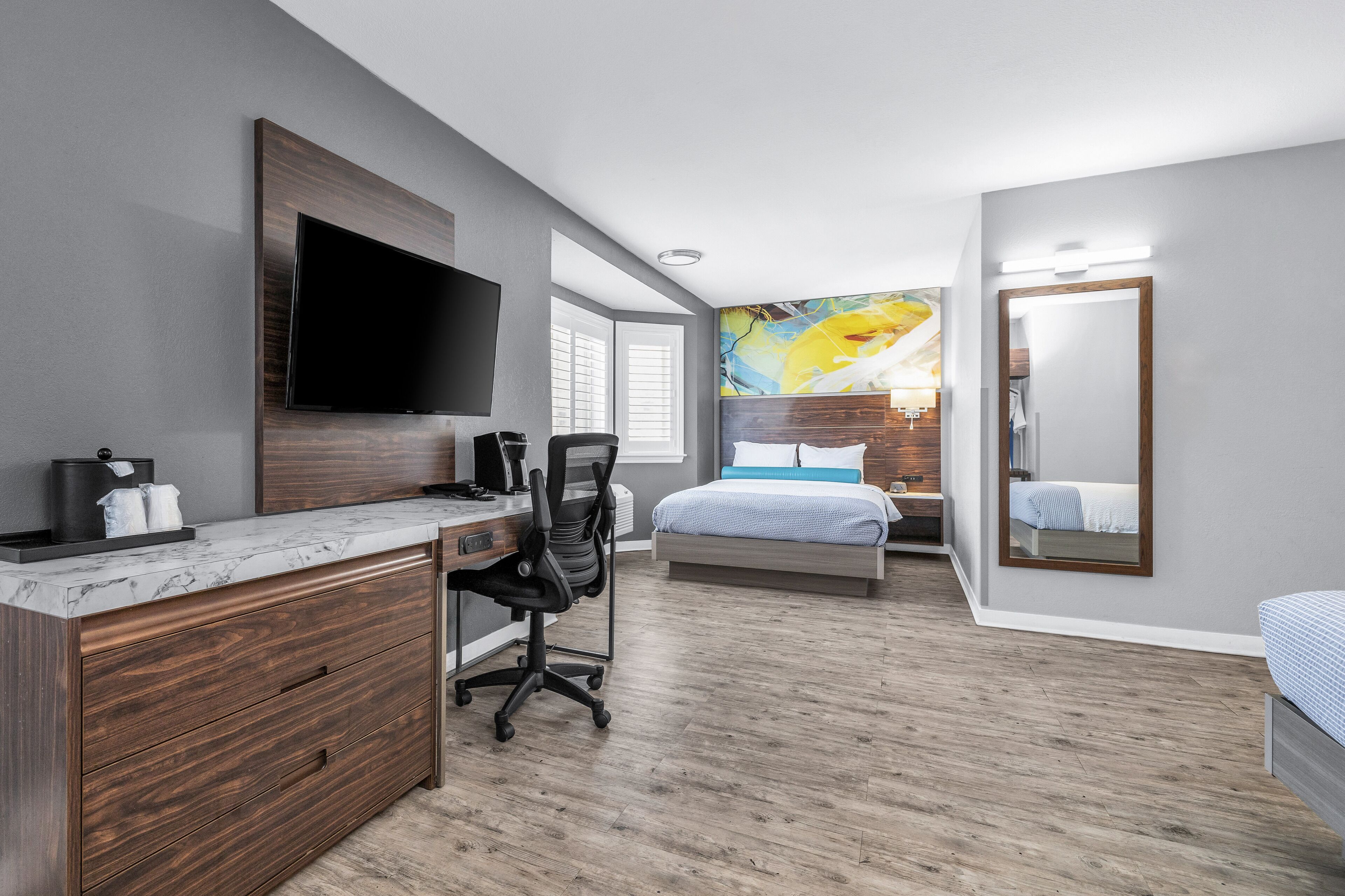 A modern hotel room with a large flat-screen TV, a wooden desk with a chair, a bed with a blue and white bedspread, and a large mirror.