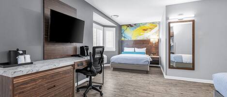 A modern hotel room with a large flat-screen TV, a wooden desk with a chair, a bed with a blue and white bedspread, and a large mirror.