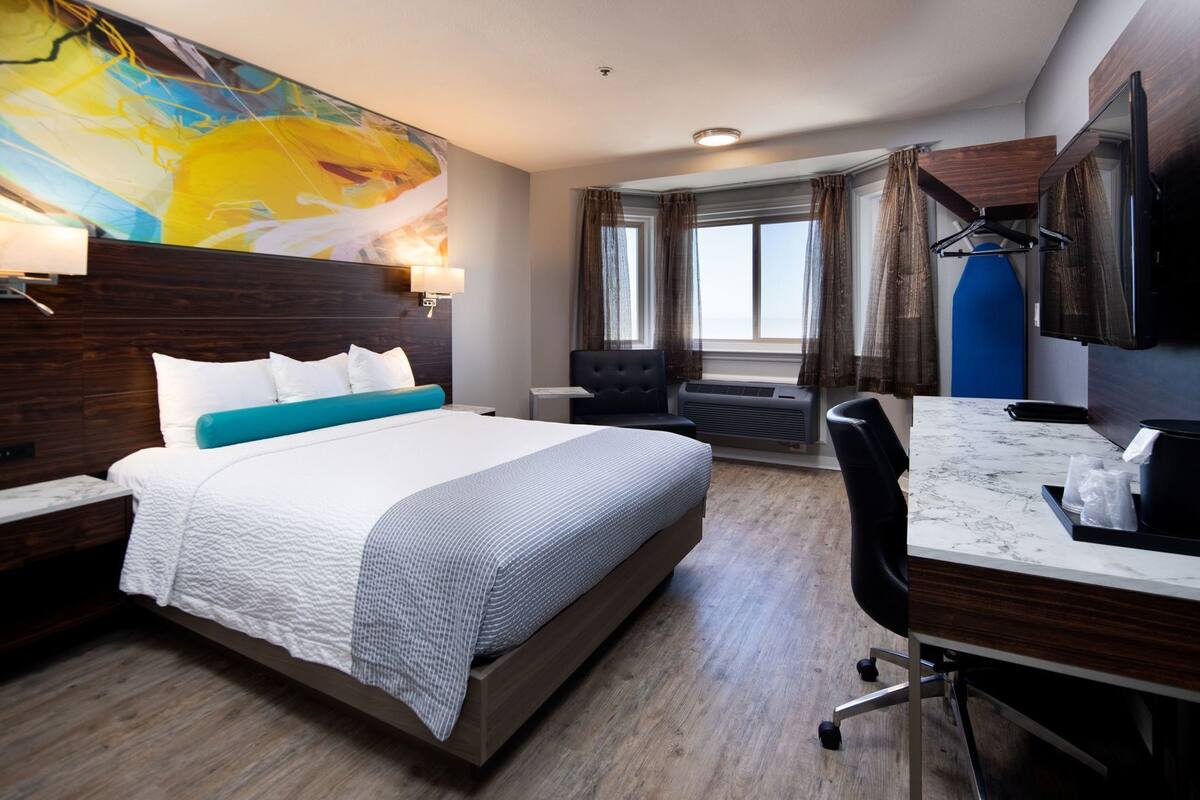 A hotel room with a large bed, a desk, a chair, a TV, and a large abstract painting on the wall.