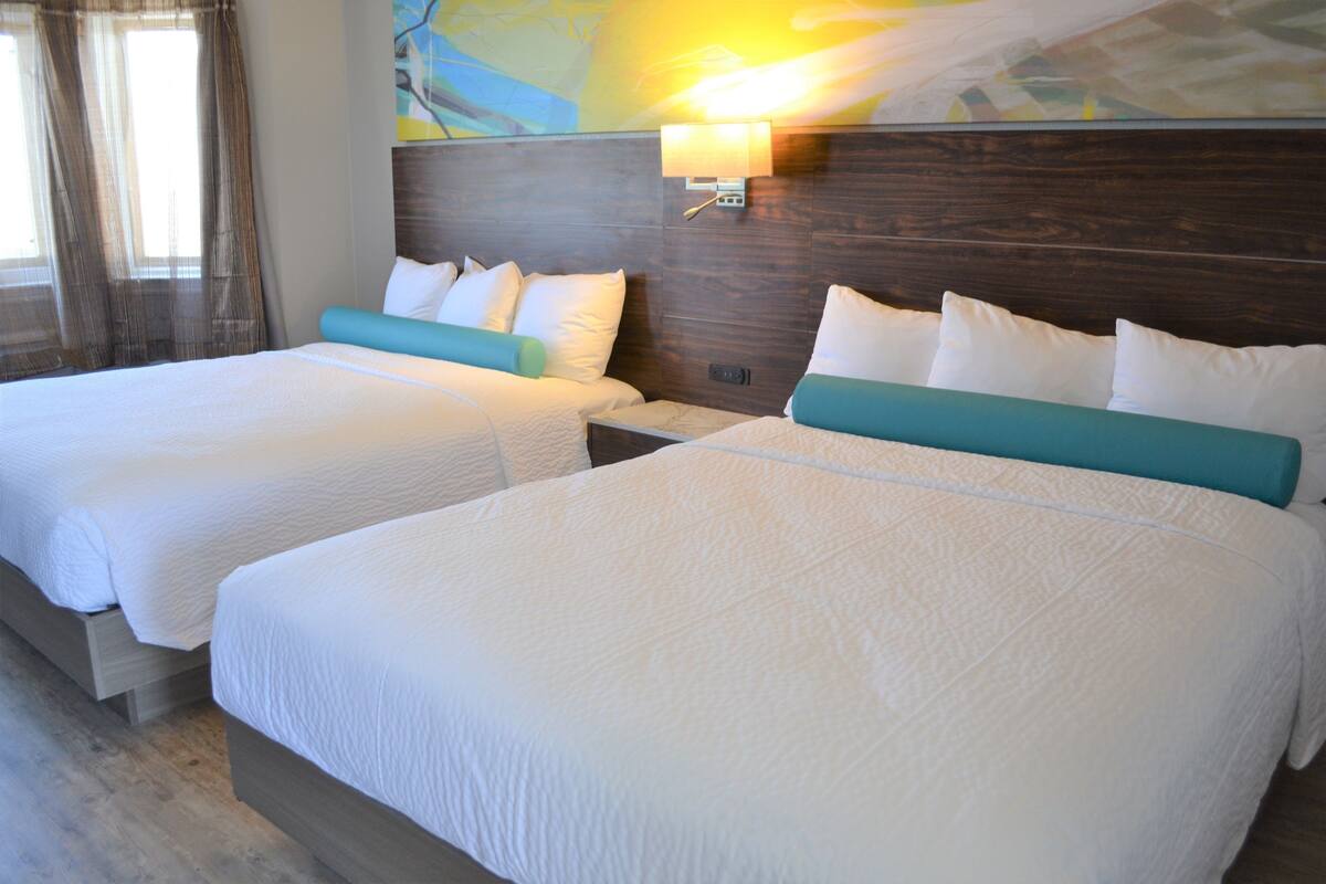 A hotel room with two beds, each with white bedding and teal headboards. There is a wall-mounted light fixture above the beds.
