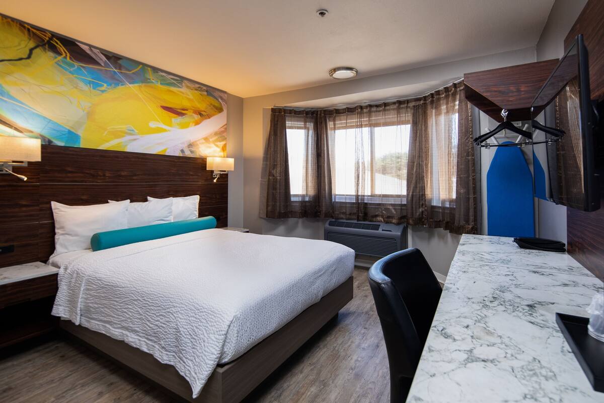 A hotel room with a bed, a desk, a chair, a TV, and a painting on the wall.