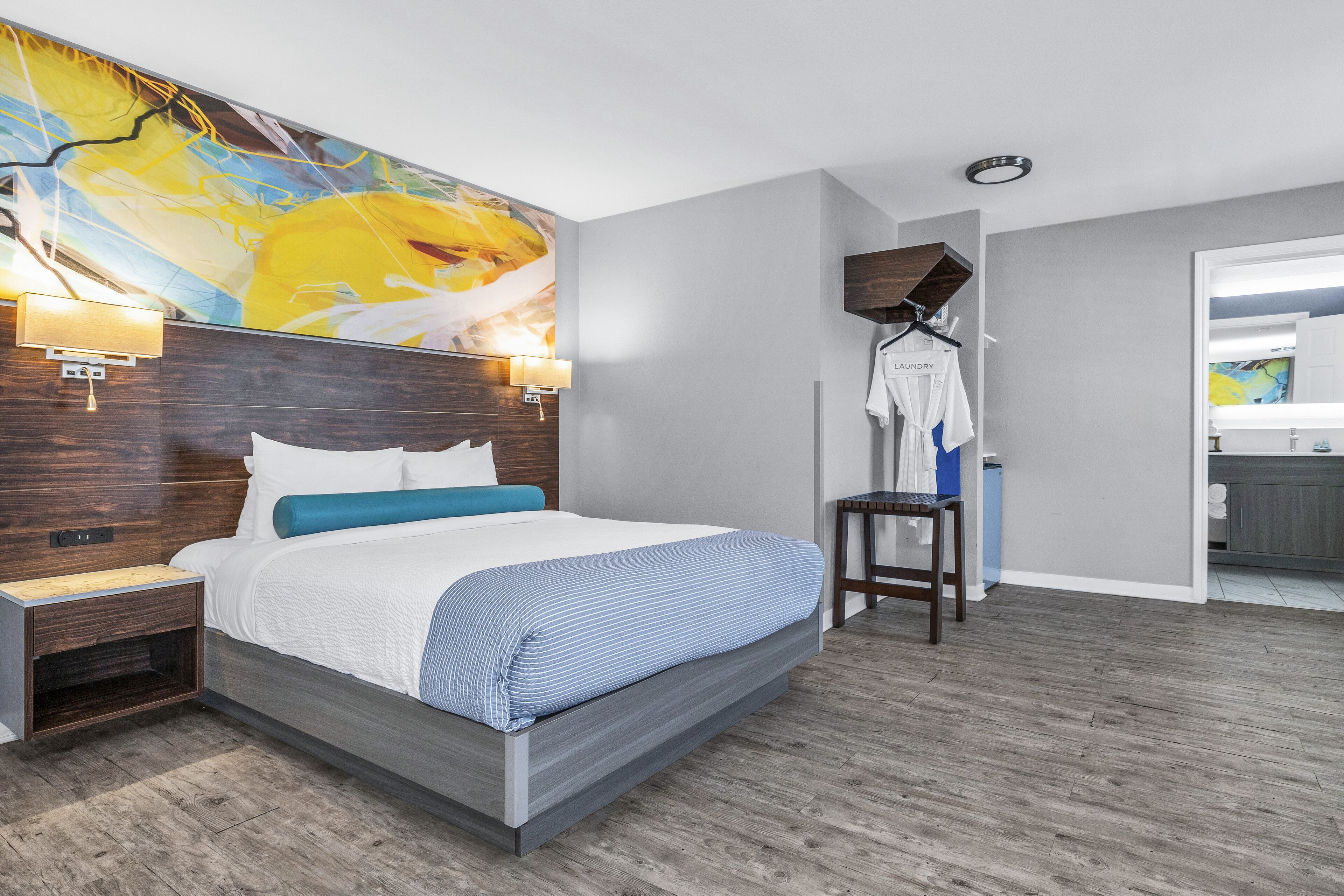 A modern hotel room with a large bed, a nightstand, a wall-mounted lamp, a hanging coat rack, and a view into the bathroom.