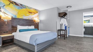 A modern hotel room with a large bed, a nightstand, a wall-mounted lamp, a hanging coat rack, and a view into the bathroom.