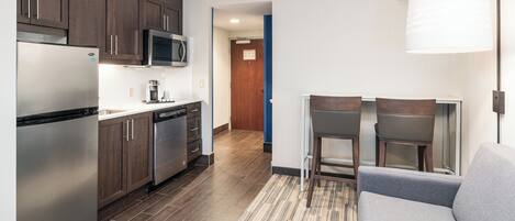 A compact kitchen with modern appliances, a dining area with a grey sofa, and a hallway leading to a door.