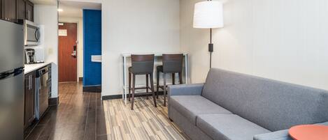 A modern hotel room with a kitchenette, a gray sofa, a small round table, and a blue accent wall.