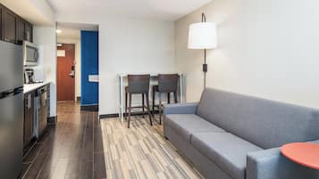 Une chambre d’hôtel moderne équipée d’une kitchenette, d’un canapé gris, de deux fauteuils, d’une petite table et d’une lampe.