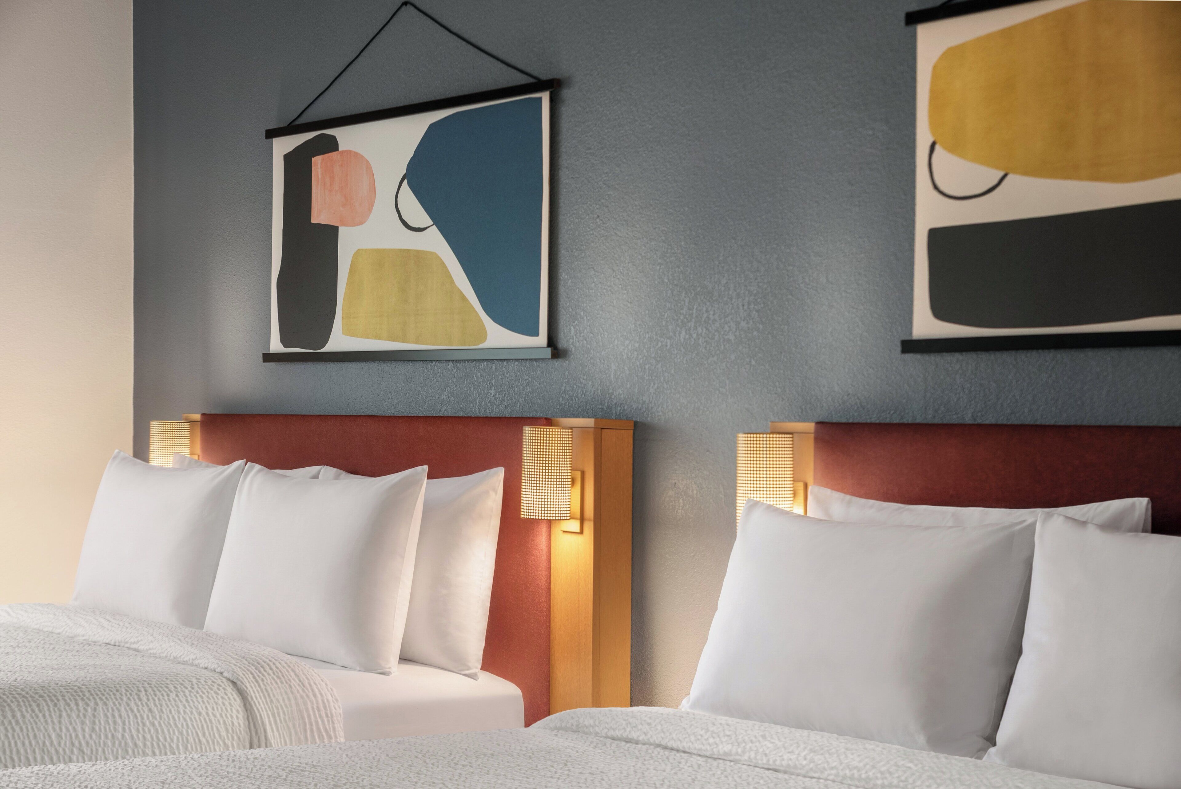 Habitación de hotel con dos camas, cada una con sábanas y almohadas blancas, cabeceros de madera y una obra de arte abstracta colgada en la pared.