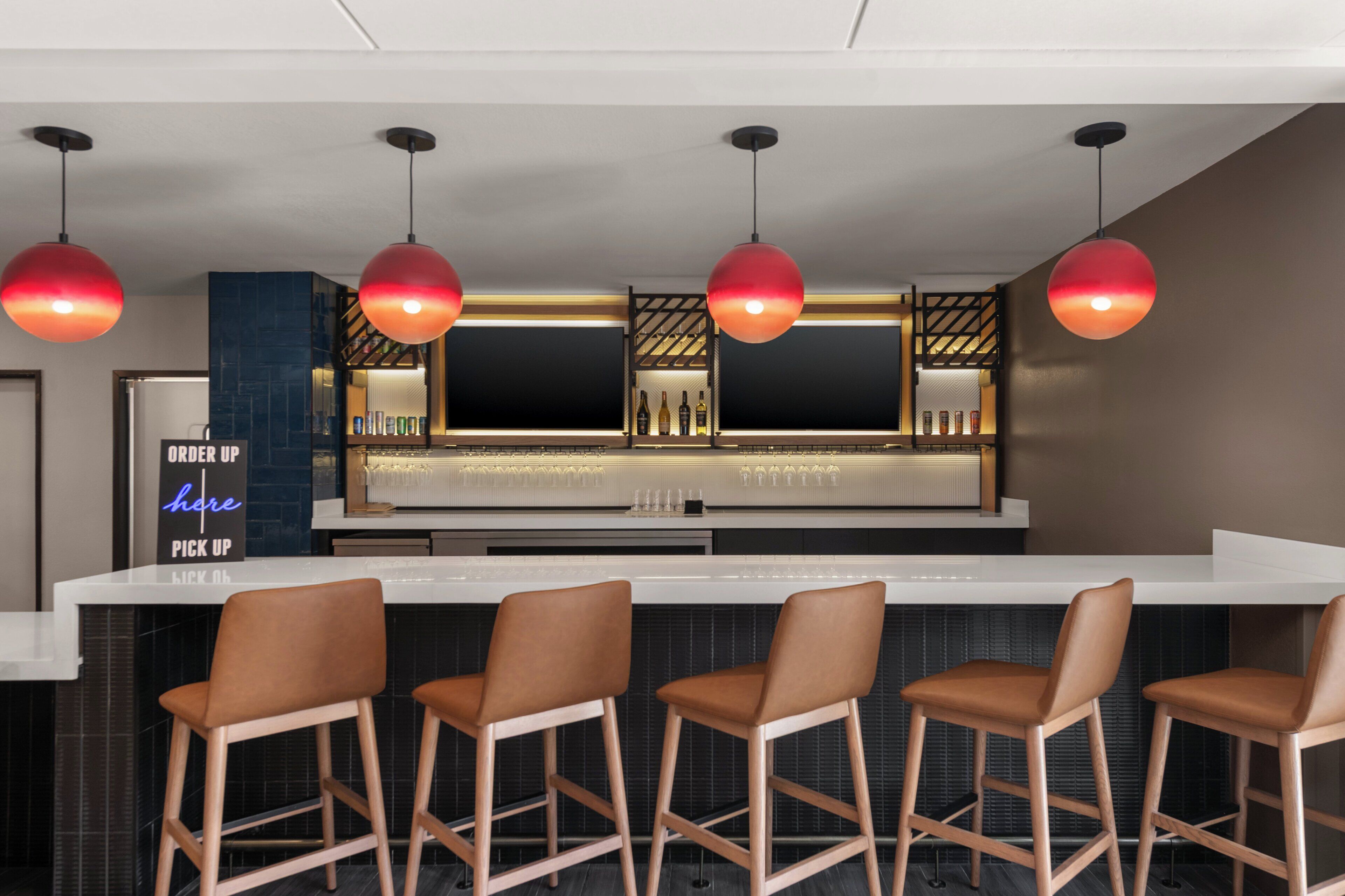 Zona de bar moderna con mostrador blanco, cuatro taburetes y lámparas colgantes rojas.