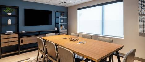 A conference room with a long table, chairs, a TV, shelves with plants, and a large window.