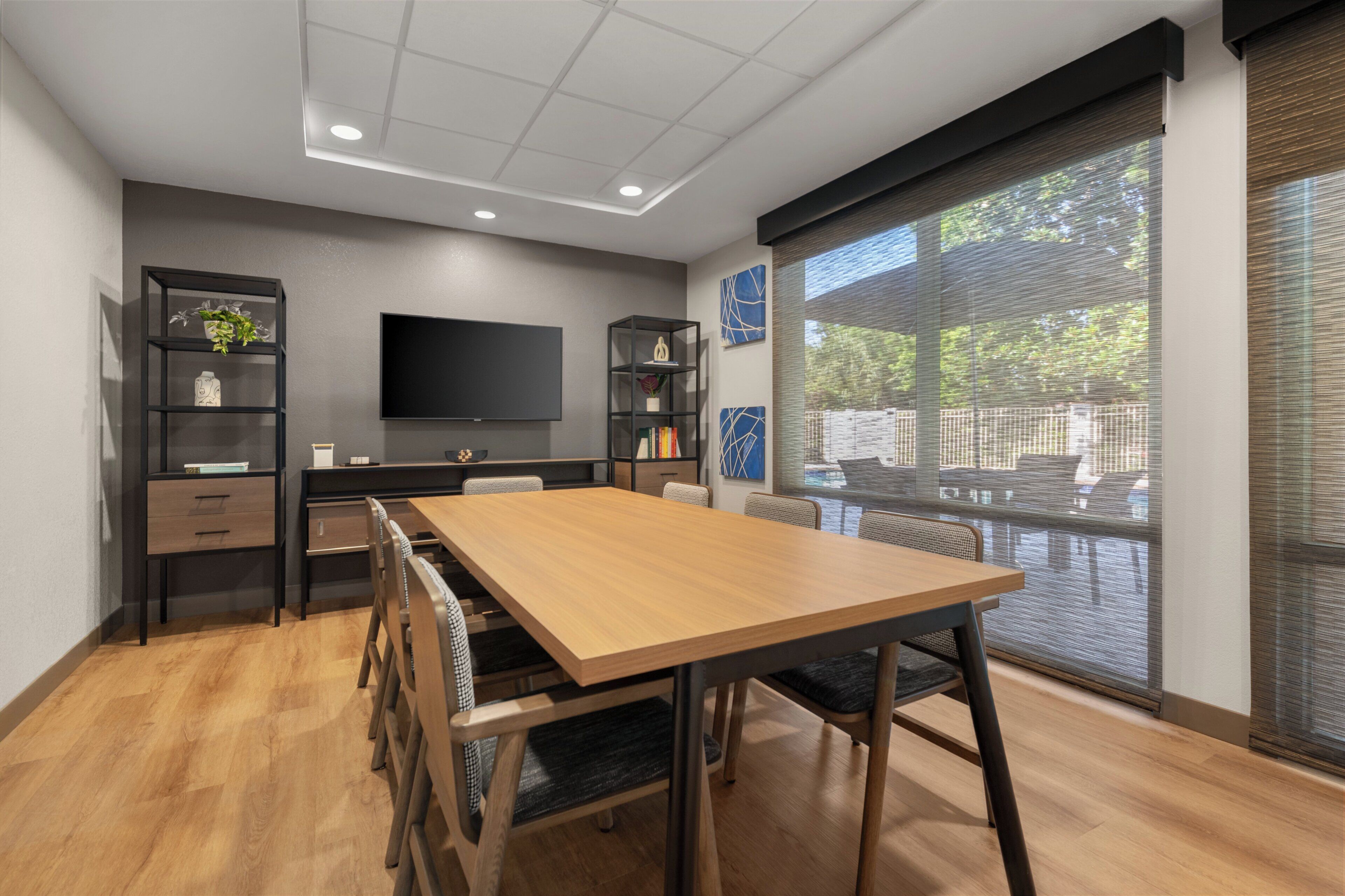 A modern dining area with a wooden table and chairs, a flat-screen TV mounted on the wall, and a large window with a view of greenery.