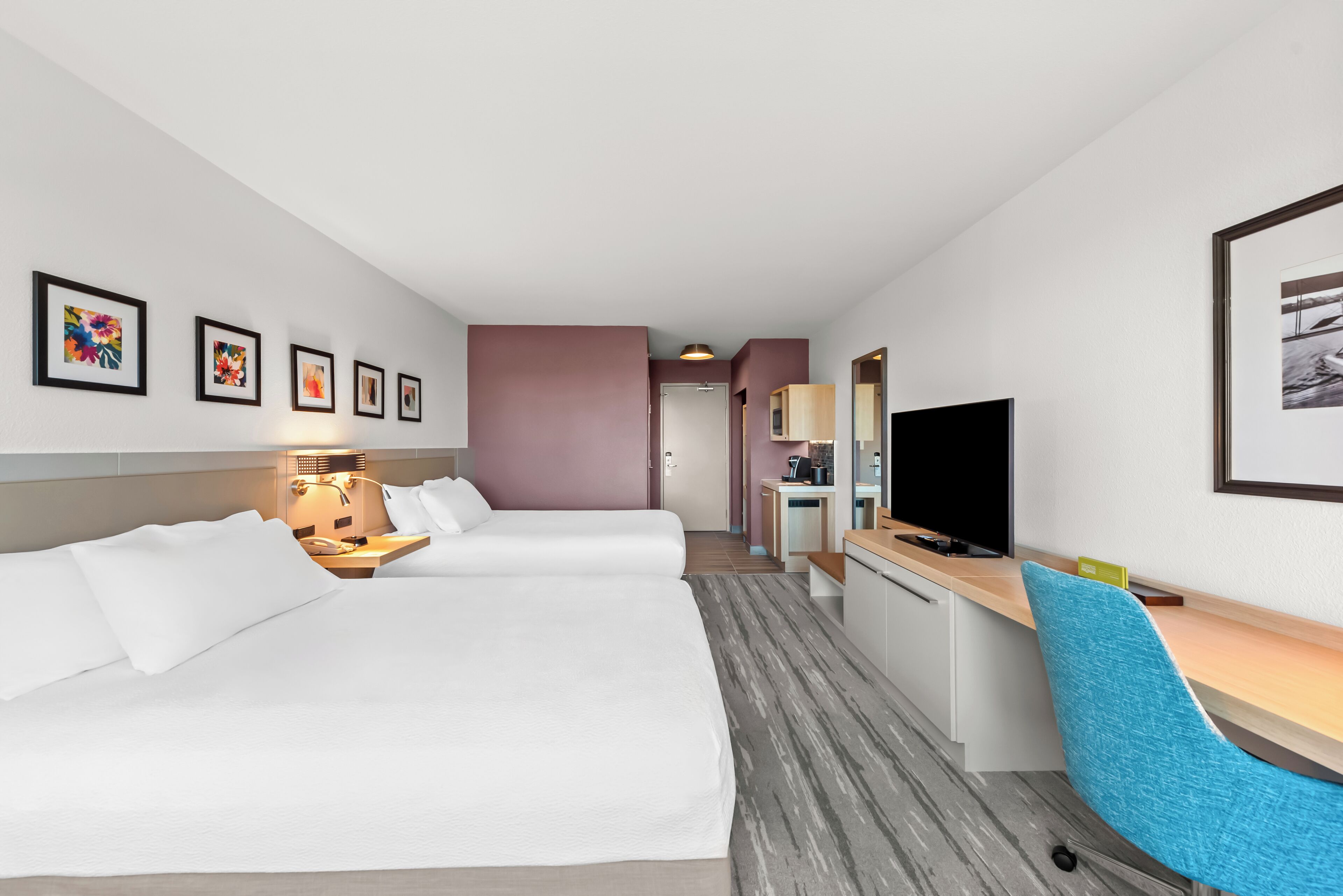 Quarto de hotel com duas camas, uma escrivaninha, uma cadeira, televisão e obras de arte nas paredes.
