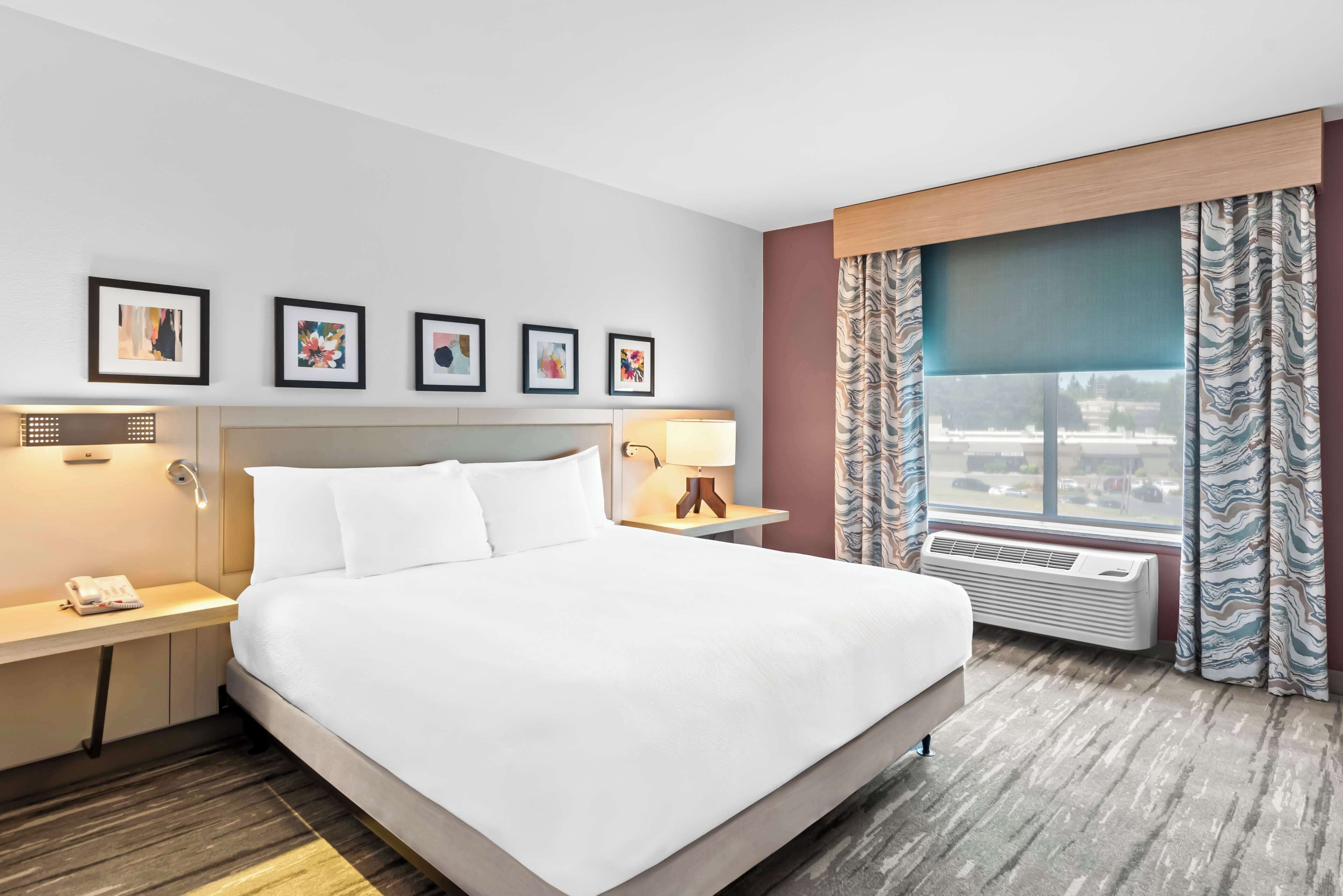 Ein Hotelzimmer mit einem großen Bett, einem Nachttisch mit Telefon, einer Lampe und einem Fenster mit Vorhängen.
