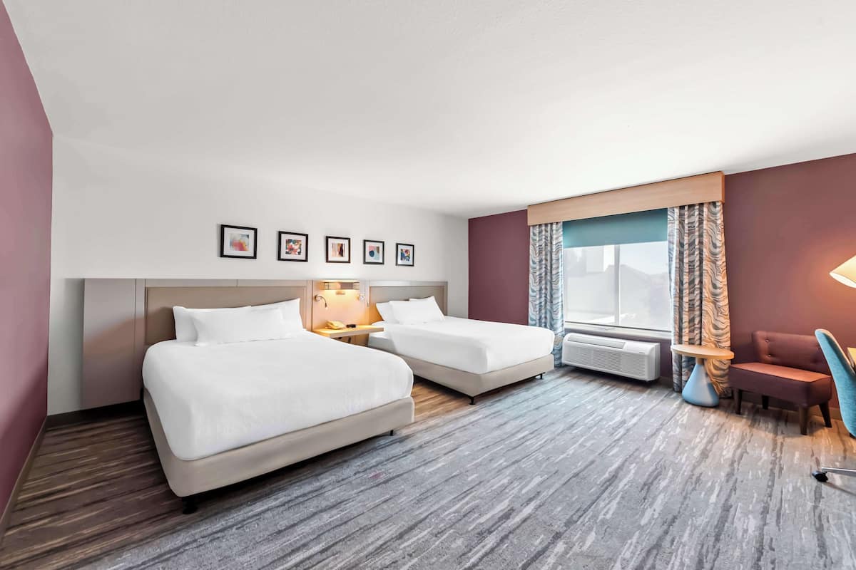 A hotel room with two beds, a desk, a chair, a window with curtains, and framed pictures on the wall.