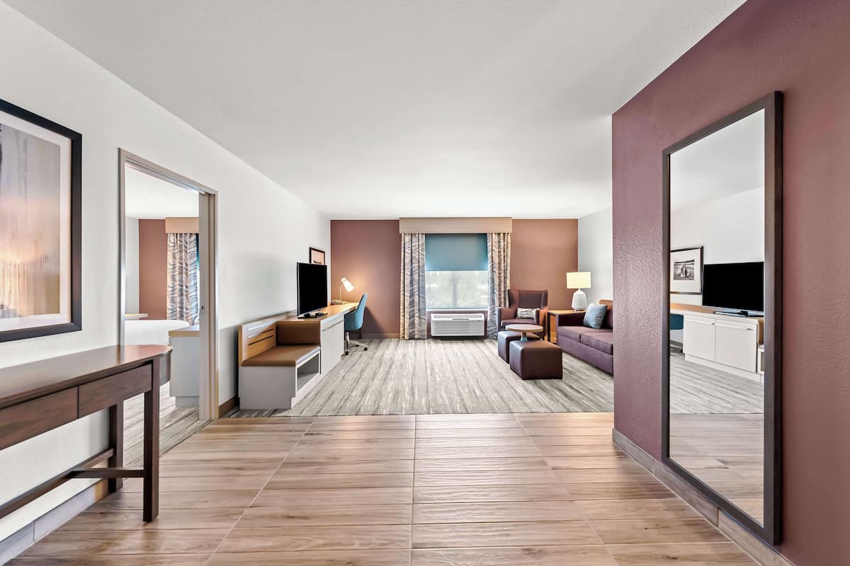 A modern hotel room with a wooden floor, a desk with a chair, a sofa, a TV, and a large mirror.
