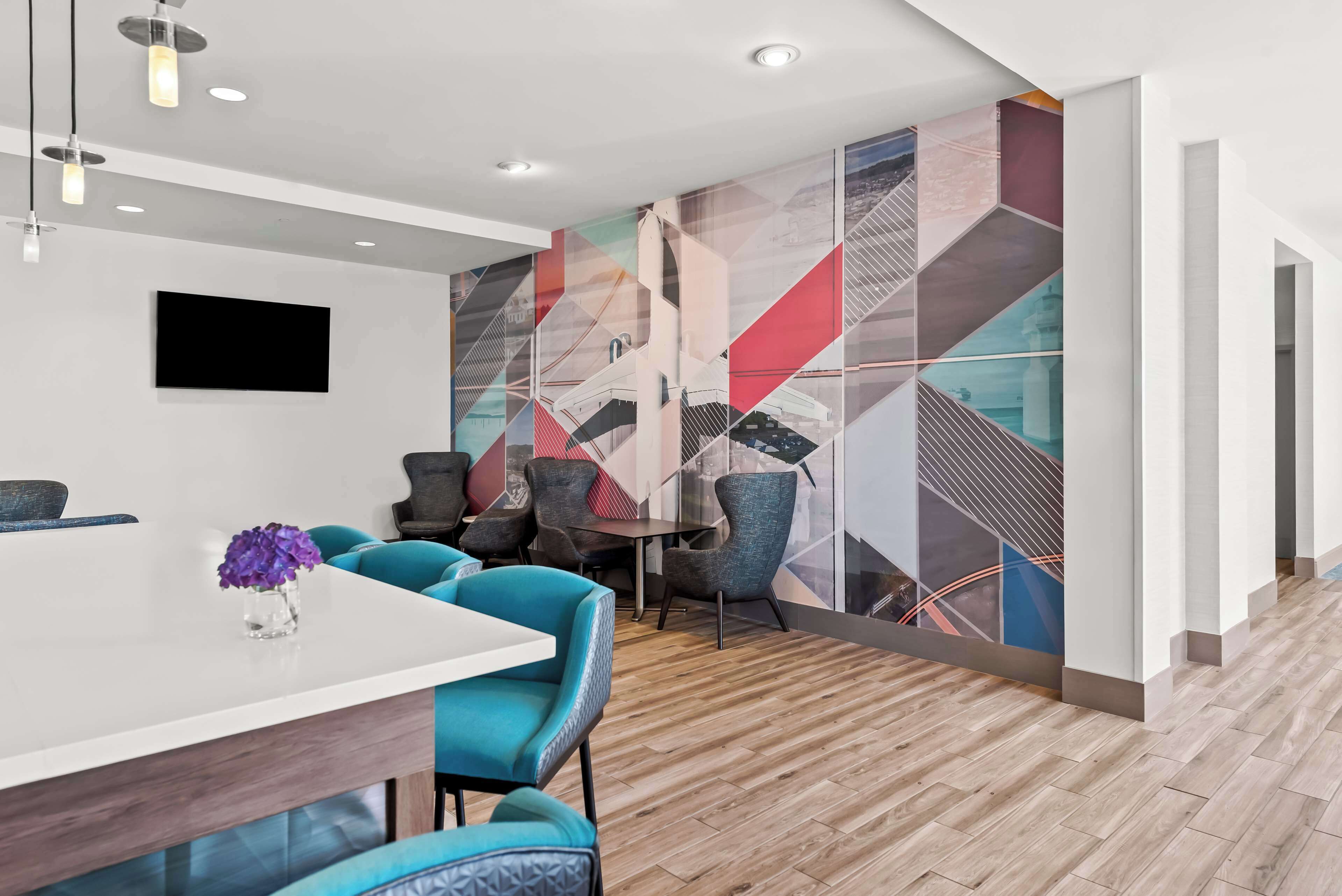 Un hall d’hôtel moderne avec un comptoir d’accueil blanc, des chaises turquoise et une fresque murale géométrique aux couleurs vives.