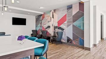 Un hall d’hôtel moderne avec un comptoir d’accueil blanc, des chaises turquoise et une fresque murale géométrique aux couleurs vives.