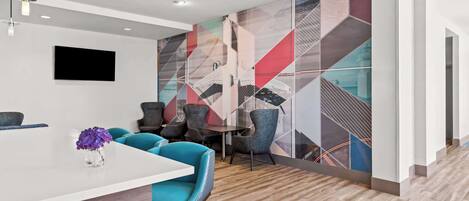 A modern hotel lobby with a white reception desk, teal chairs, and a colorful geometric wall mural.