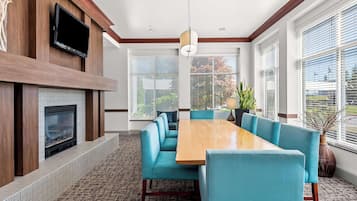 A modern dining area with a long wooden table and blue upholstered chairs, a fireplace, and large windows with blinds.