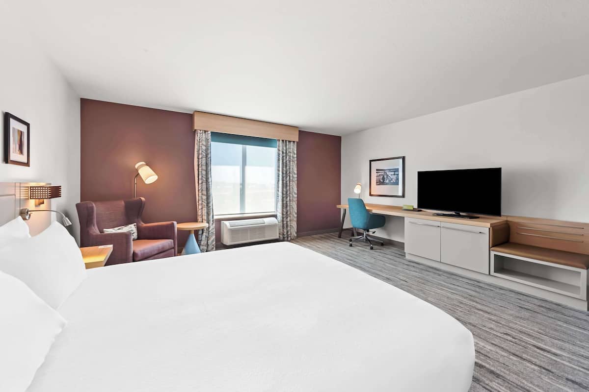 A hotel room with a large bed, a desk with a flat-screen TV, a chair, a lamp, and a window with curtains.