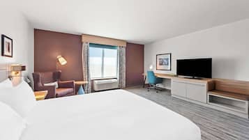 A hotel room with a large bed, a desk with a flat-screen TV, a chair, a lamp, and a window with curtains.