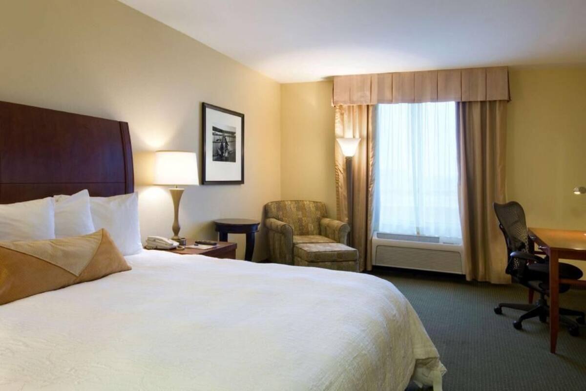 A hotel room with a large bed, a desk, a chair, a lamp, a picture on the wall, and a window with curtains.