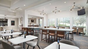 Un espace repas moderne avec des tables et des chaises blanches, une cuisine en arrière-plan et de grandes fenêtres qui laissent entrer la lumière naturelle.