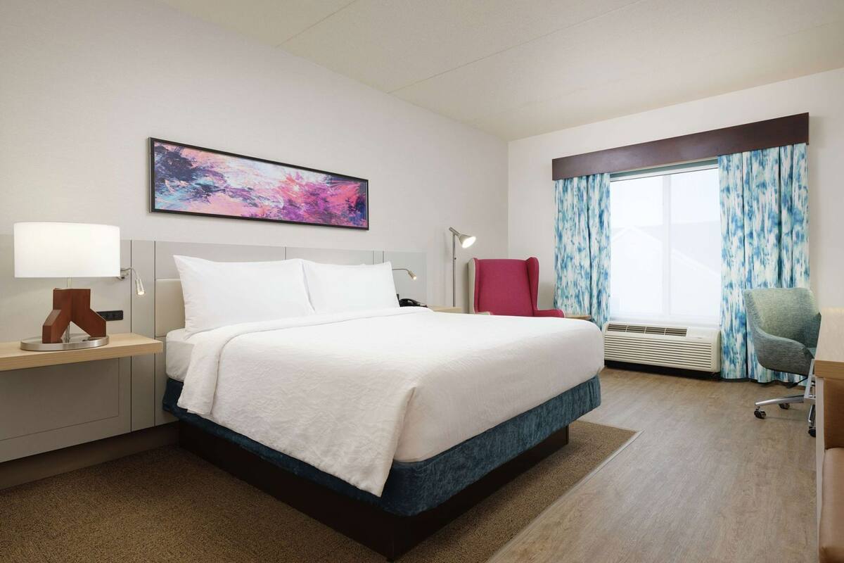 A hotel room with a bed, a desk, a chair, a window with curtains, and a painting on the wall.